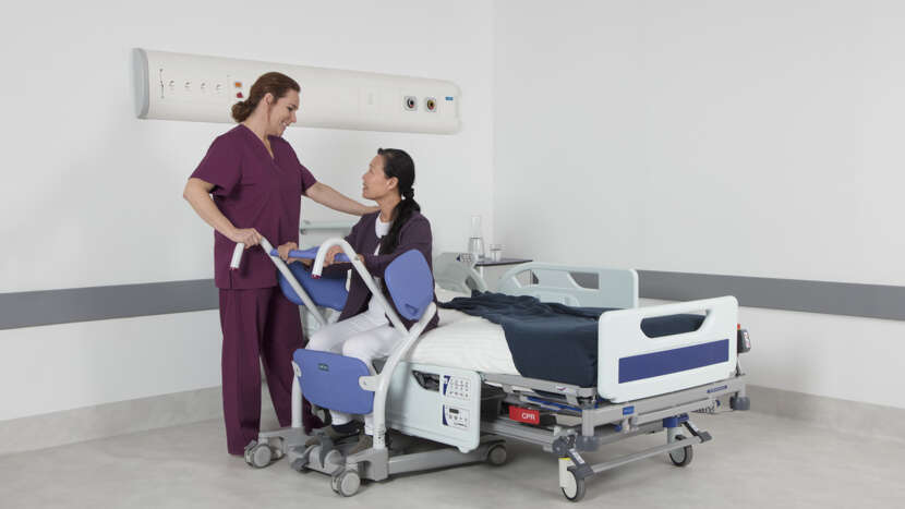 Person being helped onto a hospital bed by a nurse in a clinical setting