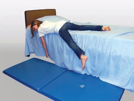 Person lying on a blue mat in front of a bed with a white sheet.