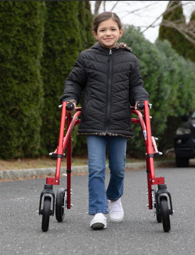 Child using a red walker on a paved path with greenery in the background