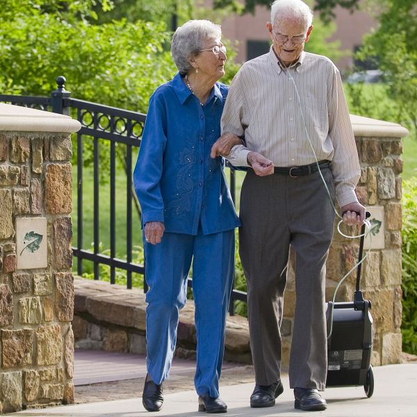 Two elderly people walking outdoors with a small device attached to the man's arm.