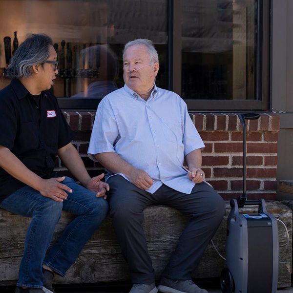 Two men sitting on a stone bench outside a building, with a dehumidifier visible.
