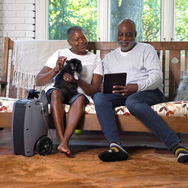 Two people sitting on a bench with a small dog, using a tablet, next to a portable oxygen concentrator.