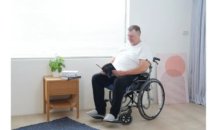 Man in a wheelchair reading a book in a room with a wooden side table and plant.
