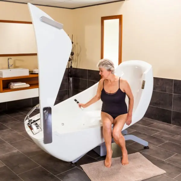 A woman in a black swimsuit sits inside a white hydrotherapy pool chair with a hinged lid, in a modern bathroom setting with dark tiled floors and walls.