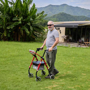 A man with a gray beard and sunglasses pushes a red and black four-wheel rolling walker with a seat and storage basket across a green lawn. A modern house with a patio and lush green mountains are in the background.