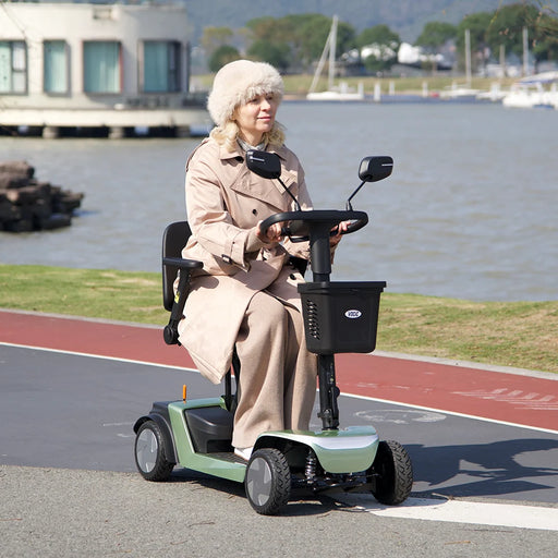Person using a mobility scooter by a waterfront with boats and houses in the background