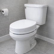 A white ceramic toilet with a closed lid and tank, shown in a bathroom setting with gray tiled walls and floor, a toilet paper holder with a roll, and white baseboards.
