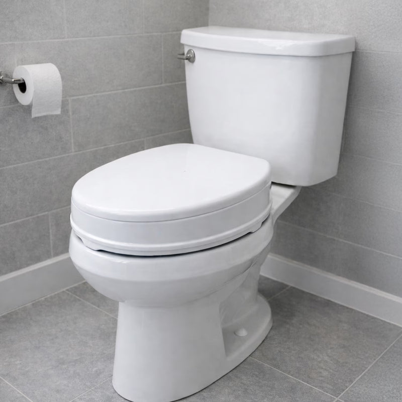 A white ceramic toilet with a closed lid and tank, shown in a bathroom setting with gray tiled walls and floor, a toilet paper holder with a roll, and white baseboards.