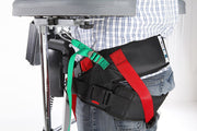 A close-up of a person wearing a black waist support belt with red and green straps, shown from the waist down next to a chair or medical support frame.