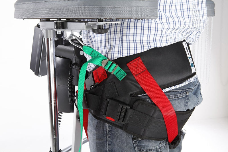 A close-up of a person wearing a black waist support belt with red and green straps, shown from the waist down next to a chair or medical support frame.