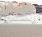 A white quilted mattress protector or comforter pad with green trim and white hanging ties, shown on a bed with a person partially visible underneath, wrapped in cream bedding.