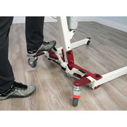 A close-up of a person's feet in black pants and gray shoes stepping onto the platform of a white and red industrial hoist or lift mechanism mounted on casters, set on a light wood floor.