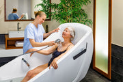 A woman relaxes in a white curved bathtub while a spa therapist showers her head with a handheld sprayer. The setting is a modern spa room with a wooden vanity, mirror, green plant, and dark tiled floor.