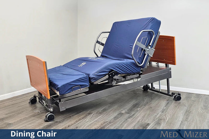 Hospital bed with blue cover and dining chair attachment on a wooden floor.