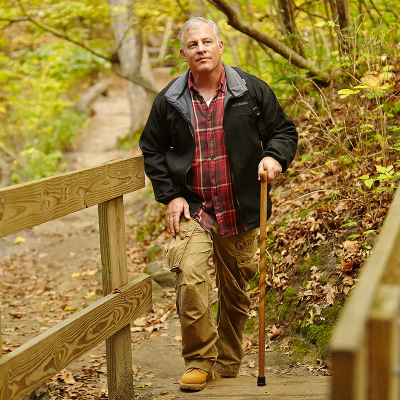 Man standing on a wooden path in a forest, holding a walking stick.