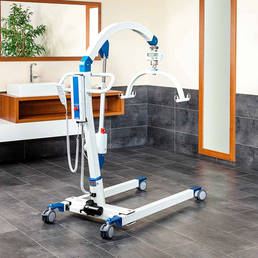 A white and blue hospital-style patient lift with wheels is shown in a bathroom setting with dark gray tile floors and walls, a sink, mirror, and plant visible.