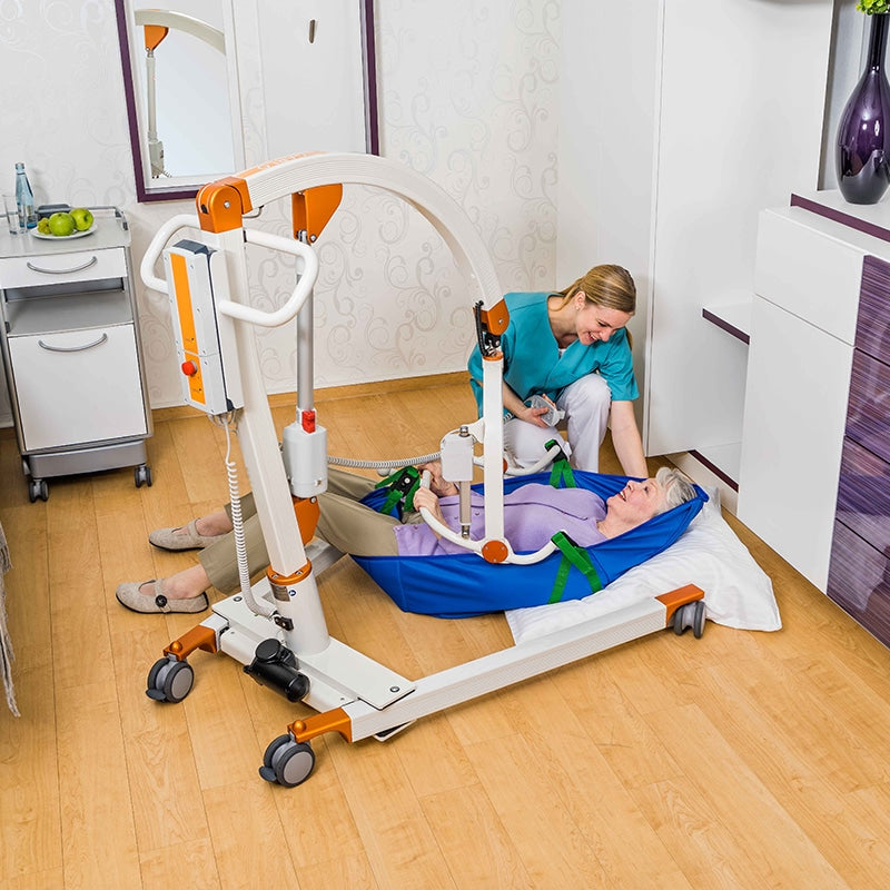 An electric patient transport bed with orange and white accents is shown in a home care setting, with a caregiver assisting a patient lying on a blue transfer pad on a wooden floor.