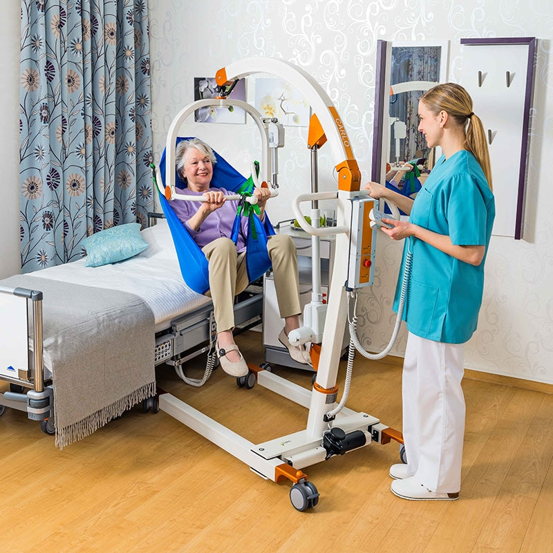 A healthcare worker in teal scrubs operates a white and orange patient transfer lift next to a hospital bed with an elderly woman sitting inside a blue sling, in a brightly lit room with patterned curtains and wood floors.