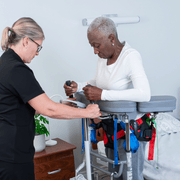 A caregiver in a black uniform assists an older woman with a mobility scooter or transfer board in a bright, clean room.