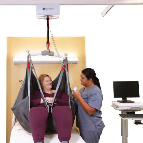 A patient lift chair with a woman sitting inside, shown in a clinical room setting with a healthcare worker standing beside it. The lift is suspended from a ceiling mount above a hospital bed.