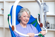 An elderly woman with short gray hair is seated in a blue and green support chair with white armrests, smiling at the camera in an indoor room with white shelves in the background.