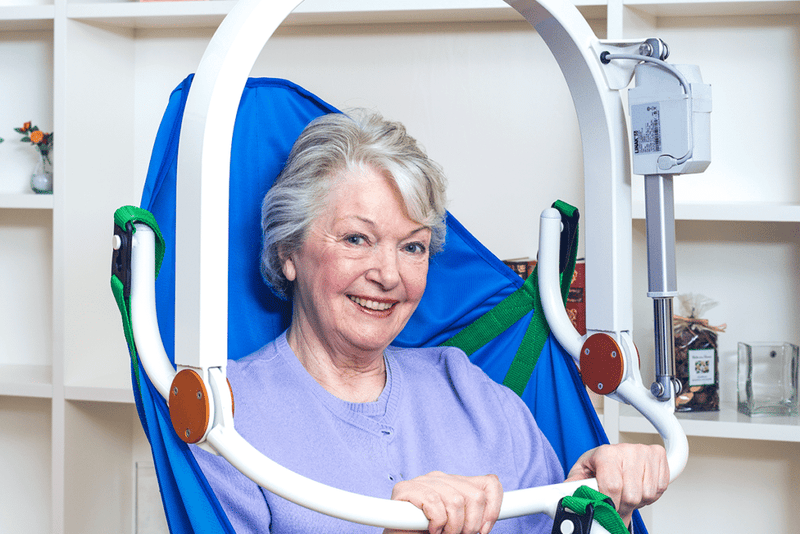 An elderly woman with short gray hair is seated in a blue and green support chair with white armrests, smiling at the camera in an indoor room with white shelves in the background.