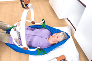 A person reclining in a blue manual wheelchair lift sling with a white frame and green straps on a wooden floor.