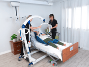 A medical transport bed with a patient in a blue sling being assisted by two caregivers in a bright, modern bedroom setting.