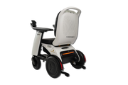 A modern power wheelchair shown from a three-quarters rear angle on a solid white background. The wheelchair features a white and black frame, a high backrest with a textured back panel, black wheels, and a small orange LED light indicator on the rear.