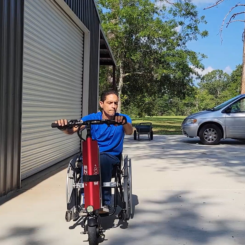 Person using a red and black mobility scooter in a driveway with a storage unit and car in the background.