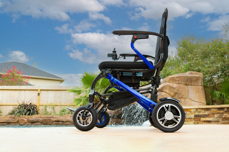 A blue and black electric wheelchair with large rear wheels and smaller front casters, photographed outdoors beside a pool with a house, trees, and sky in the background.