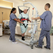 An elderly man in a hospital gown and socks is being assisted by two nurses using a hospital bed transfer lift in a clinical room.
