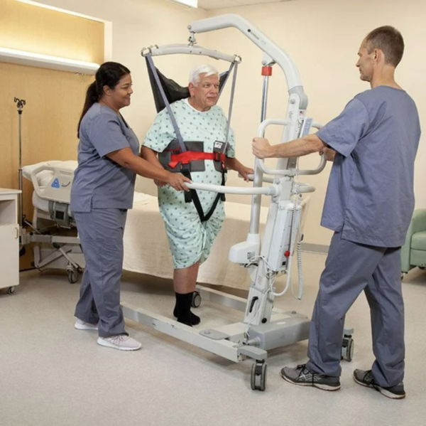 An elderly man in a hospital gown and socks is being assisted by two nurses using a hospital bed transfer lift in a clinical room.