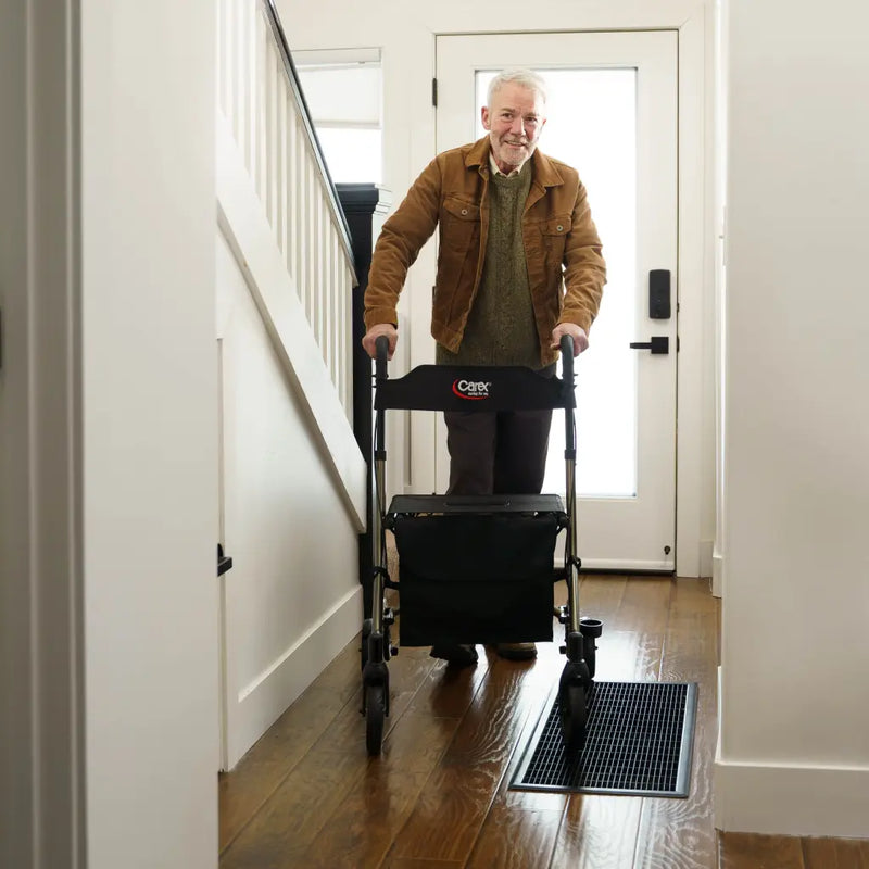 An older man stands in a hallway using a black Cargotrol rolling luggage cart with a storage bag, on a hardwood floor.