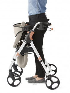 A white and black four-wheel rolling walker with a beige storage basket, shown being used by a person wearing black pants and gray slip-on shoes against a white background.