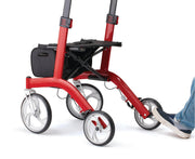 A red and black four-wheel rolling walker with a black storage basket, shown on a white background with a person's foot pushing one of the wheels.