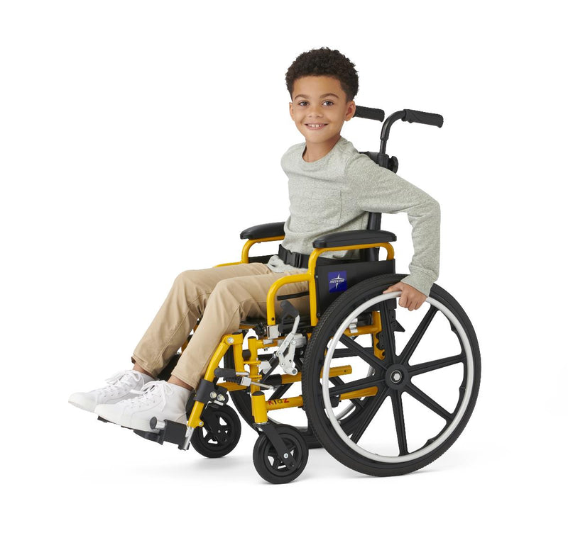 A young boy smiling while sitting in a yellow and black manual wheelchair, wearing a light gray sweater, beige pants, and white sneakers, photographed against a solid white background.