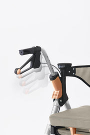 Close-up of a rollator handlebar assembly with black grips, silver metal tubing, and a beige fabric seat against a clean white background.