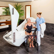 A white electric shower chair with a built-in seat and armrests is shown in a bathroom setting with a woman sitting inside and a caregiver standing beside her using a walker.