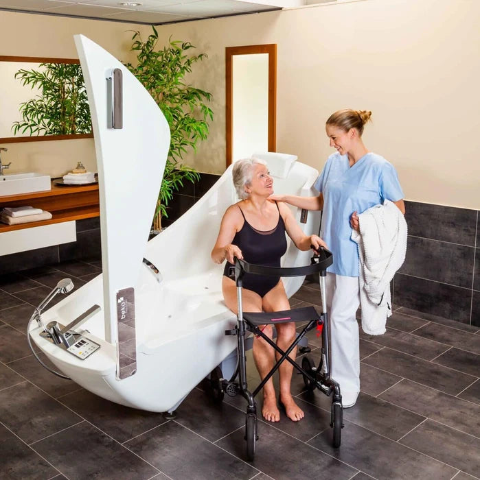 A white electric shower chair with a built-in seat and armrests is shown in a bathroom setting with a woman sitting inside and a caregiver standing beside her using a walker.
