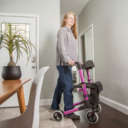 A woman with glasses stands indoors next to a bright pink and black rolling walker, wearing a gray button-up shirt and blue jeans. The setting is a home hallway with gray walls, a wooden floor, a potted plant, and a dining table in the foreground.