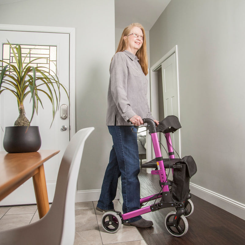 A woman with glasses stands indoors next to a bright pink and black rolling walker, wearing a gray button-up shirt and blue jeans. The setting is a home hallway with gray walls, a wooden floor, a potted plant, and a dining table in the foreground.