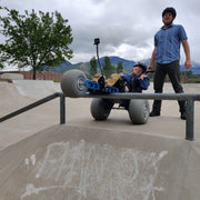 Person using a wheelchair with large wheels at a skate park, assisted by another person.