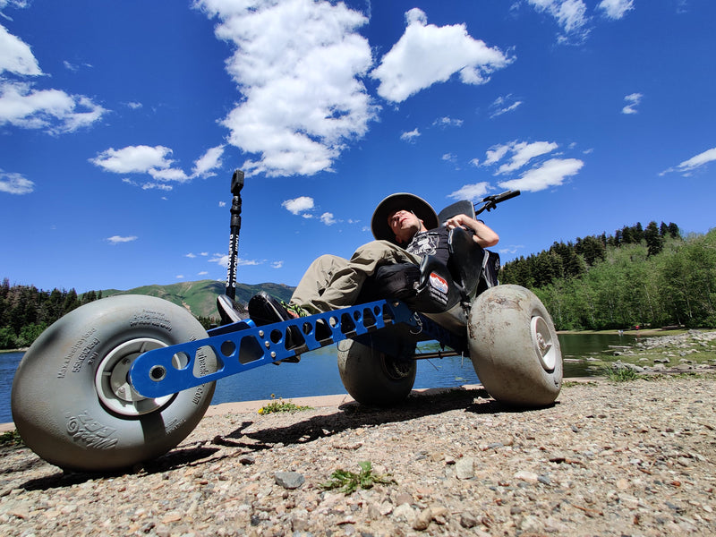 Person sitting on a large wheeled vehicle by a lake with mountains in the background