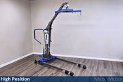 A high-position medical lift chair with a blue base and arm, gray frame, and black wheels, shown in a room corner with gray walls and wood-look flooring. A blue banner at the bottom reads 'High Position' and 'MED MIZER'.