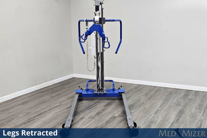 A blue and silver medical lift with a wheeled base stands in a room corner with gray wood-look flooring and light gray walls. The lift is shown with its legs retracted, and text overlays at the bottom read 'Legs Retracted' and 'MED MIZER'.