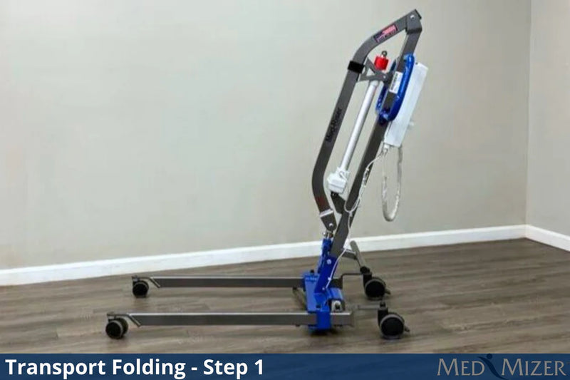 A medical transport lift in the folded position, shown on a wood-look floor against a beige wall. The product has a dark gray metal frame, blue hydraulic components, and black wheels.