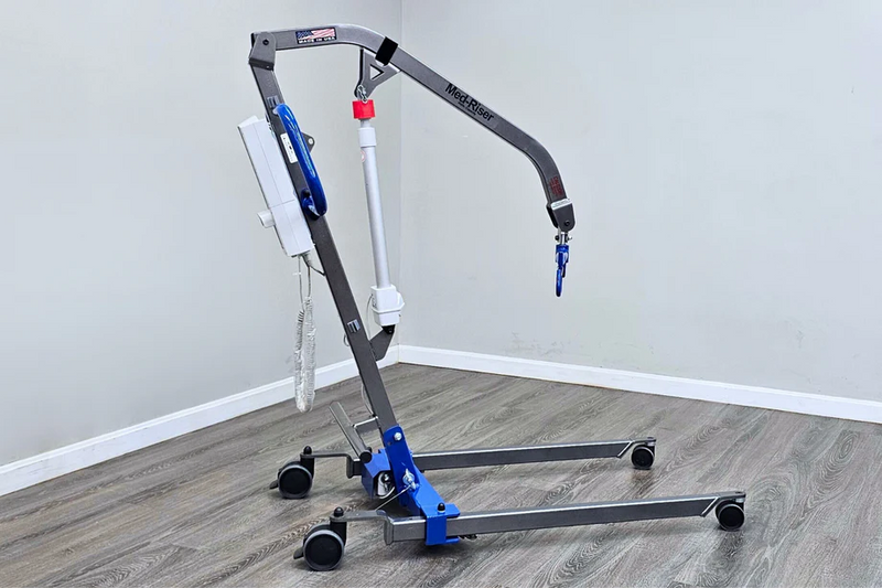 A medical lift chair with a gray metal frame, blue and white components, and black caster wheels, shown in a room with gray walls and wood-look flooring.