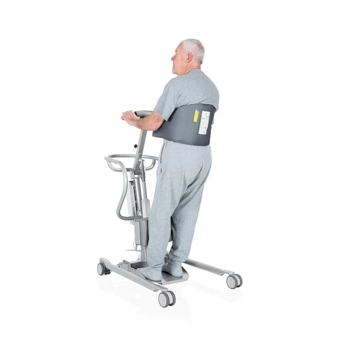 An older man wearing a gray shirt and sweatpants stands on a gray motorized transfer chair with wheels, demonstrating use of the chair. The man is shown from the back and side, with a dark gray waist support belt visible around his waist.