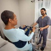 A home care or therapy setting shows a woman seated in a dark blue lifting sling attached to a transfer frame, assisted by a male therapist in gray scrubs standing beside her.